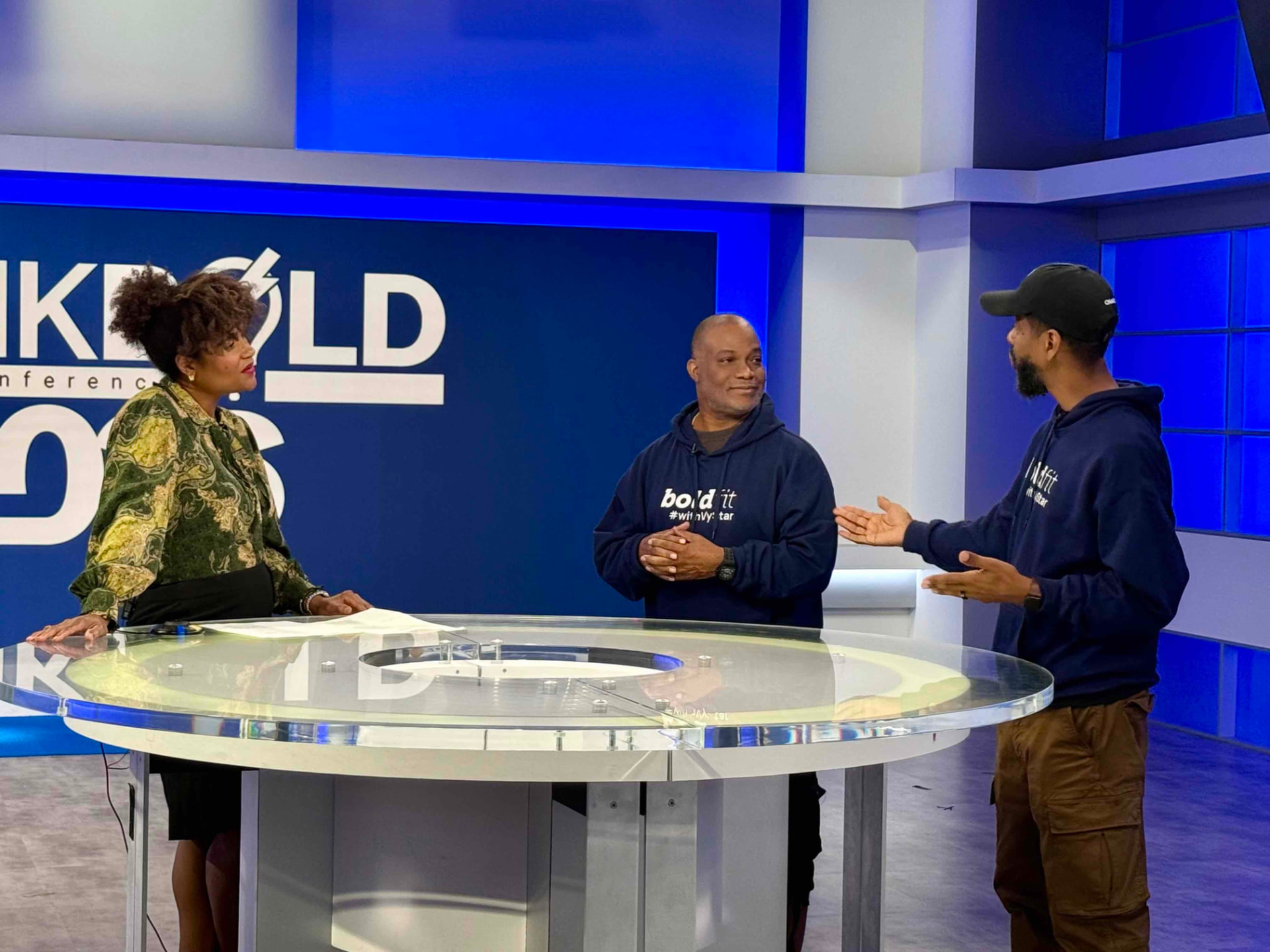Troy and Mo in Bold It hoodies talking with the host across the round studio table